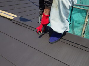 棟板金の取り付けを進めて行きます。 棟板金がきちんと真っ直ぐになるよう微調整をしながら、スクリュー釘を打ち込んでいきました。 この時に、ケミカル面戸と棟板金の隙間が開いてしまっていると雨水侵入の原因になるので、棟板金を抑えながらスクリュー釘を打ち込んで固定していきました。